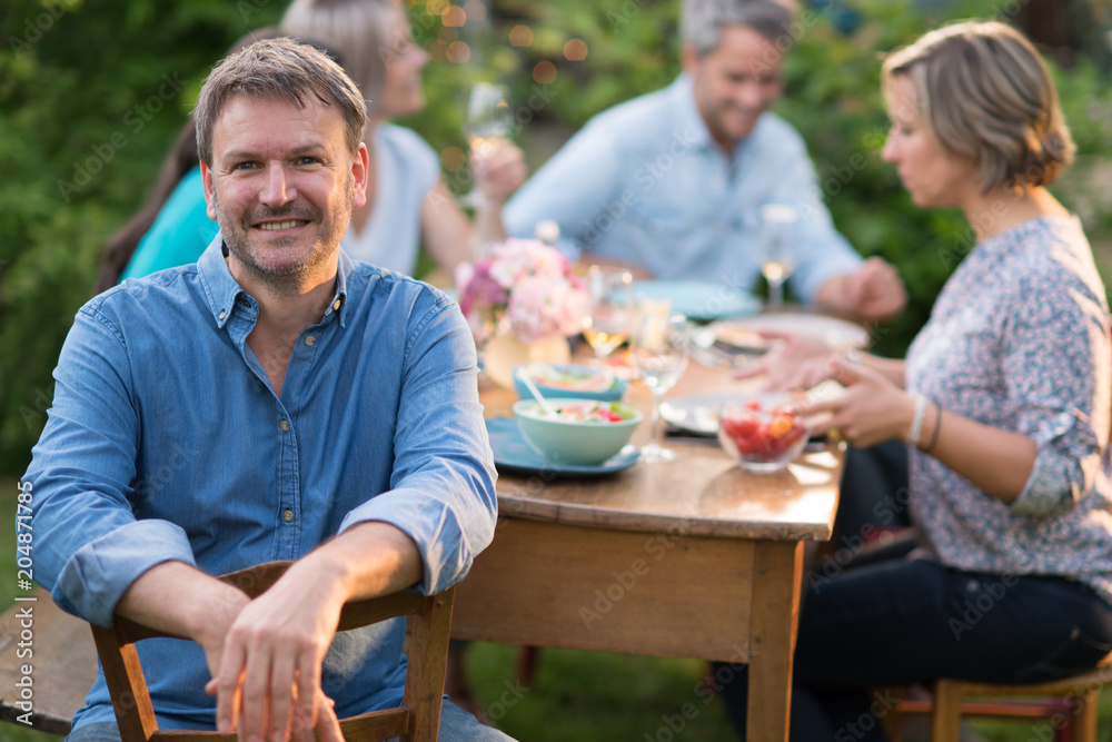 Looking at the camera, a man in his forties with a glass of wine in his hand sits at a table in the garden with his friends for dinner. Looking at the camera, a man in his forties with a glass of wine in his hand sits at a table in the garden with his friends for dinner.