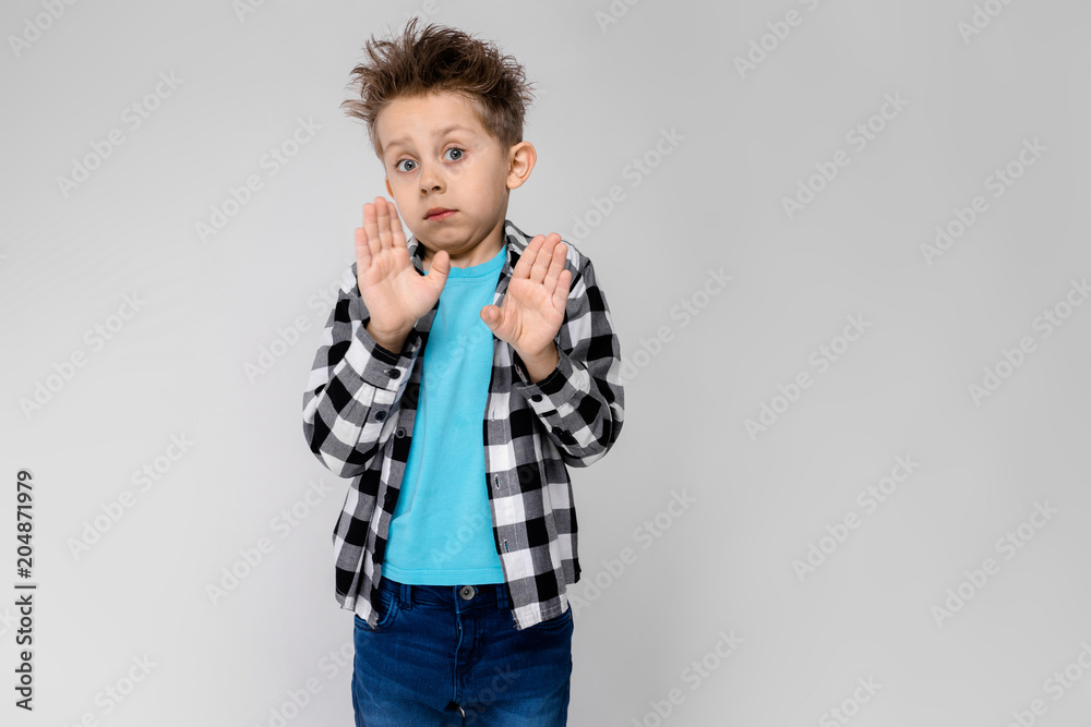 A handsome boy in a plaid shirt, blue shirt and jeans stands on a gray background. The boy folded his arms over his chest. The boy shows two palms A handsome boy in a plaid shirt, blue shirt and jeans stands on a gray background. The boy folded his arms over his chest. The boy shows two palms