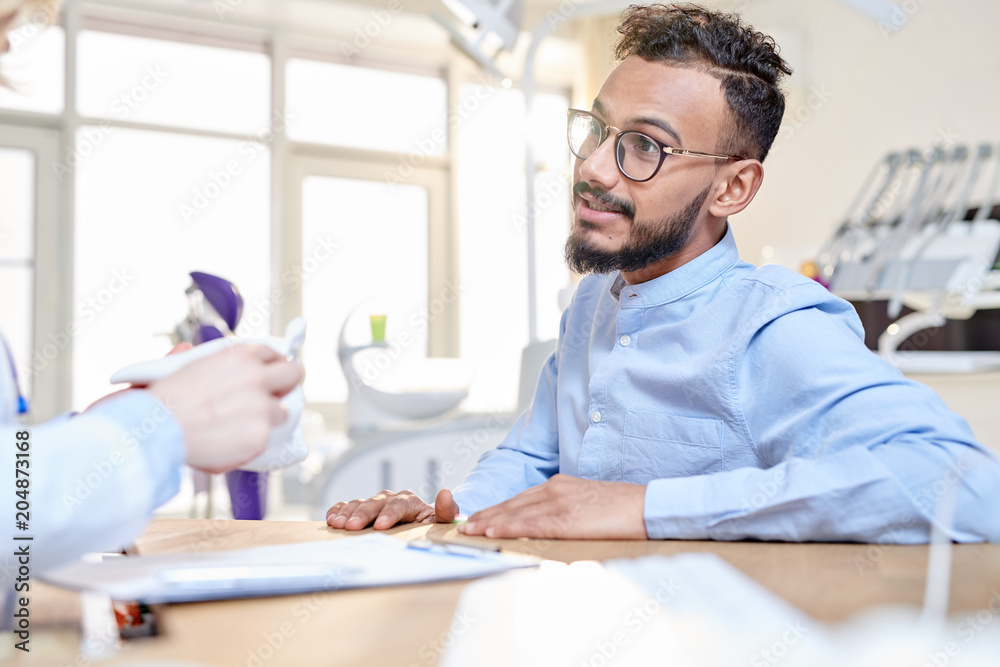 Portrait of modern Middle-Eastern man talking to dentist sitting at desk in modern dental clinic, copy space Portrait of modern Middle-Eastern man talking to dentist sitting at desk in modern dental clinic, copy space