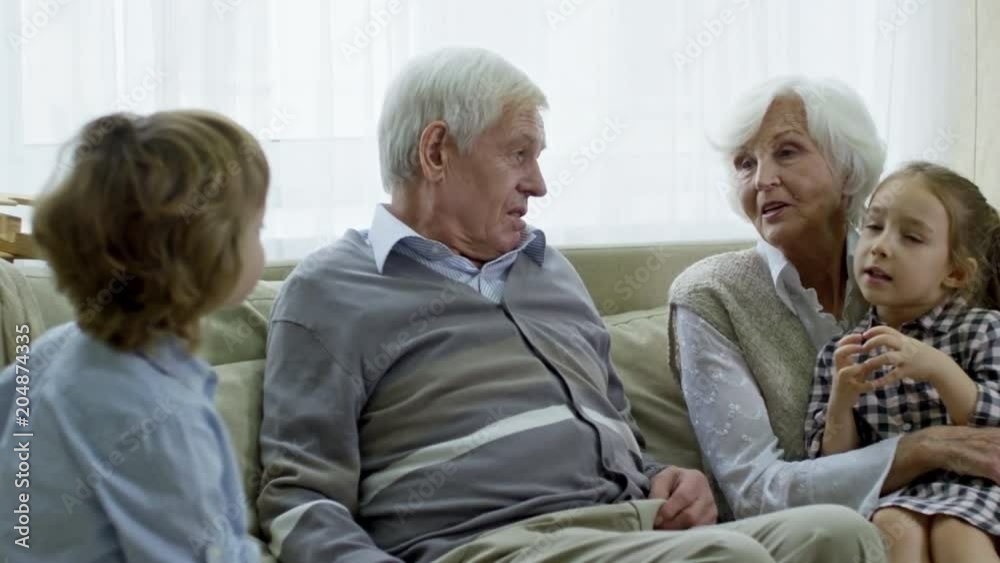 Medium shot with PAN of cheerful grandfather and grandmother with grey hair sitting on sofa and chatting with children Medium shot with PAN of cheerful grandfather and grandmother with grey hair sitting on sofa and chatting with children