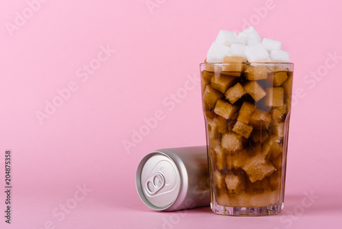 Drinking glass of of lump sugar cubes and soft cola drink with aluminum can on pink pastel background. Unhealthly diet with sweet sugary soft drinks concept.