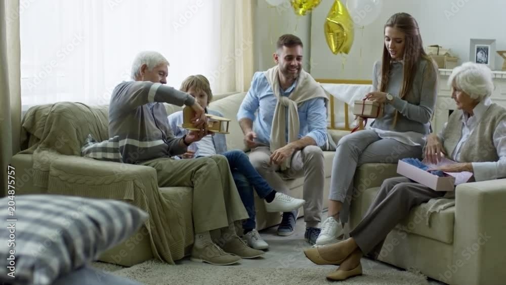 Wide shot with PAN of cheerful grandfather playing with grandson while happy young woman and grandmother opening presents on baby shower Wide shot with PAN of cheerful grandfather playing with grandson while happy young woman and grandmother opening presents on baby shower