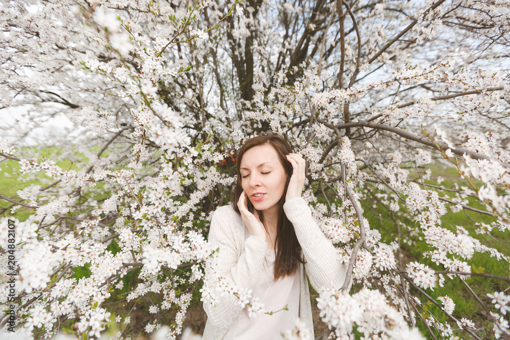 Young relaxed tender beautiful woman in light casual clothes keeping hands near face standing in city garden or park on blooming tree background. Spring nature, flowers. Lifestyle, leisure concept. Young relaxed tender beautiful woman in light casual clothes keeping hands near face standing in city garden or park on blooming tree background. Spring nature, flowers. Lifestyle, leisure concept.