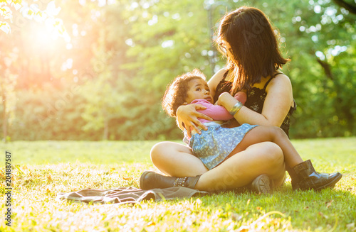 Young hispanic mother breastfeeding a dark-skinned mixed race toddler three year old daughter sitting on the green grass in nature - maternity concept. Little hispanic girl looking up. Copy space