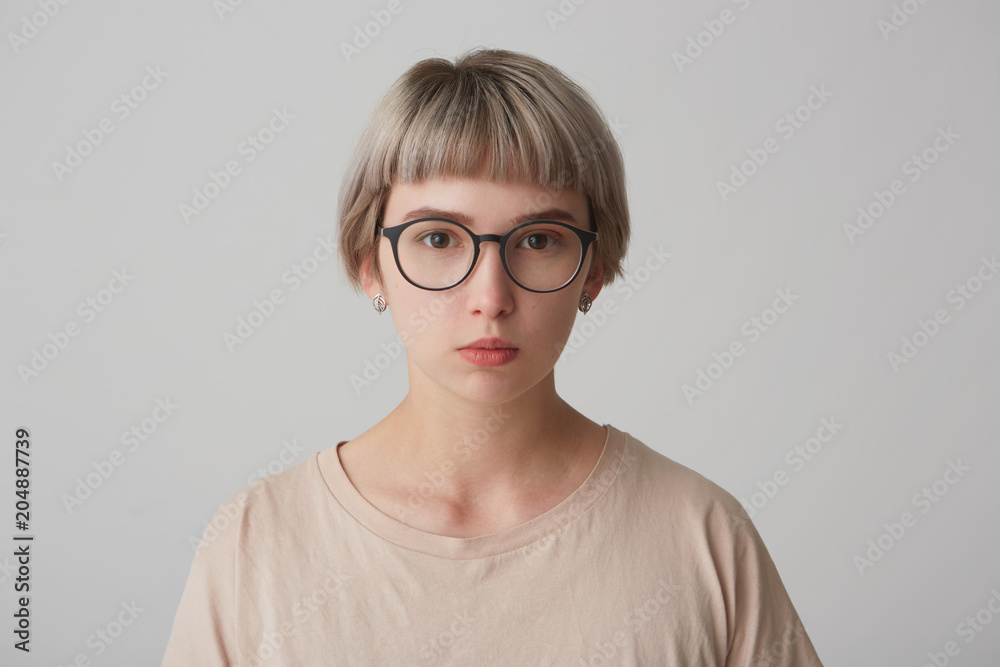 Serious female student, wears round spectacles and t shirt, looks self assured, sad and tired. Isolated over white background.
