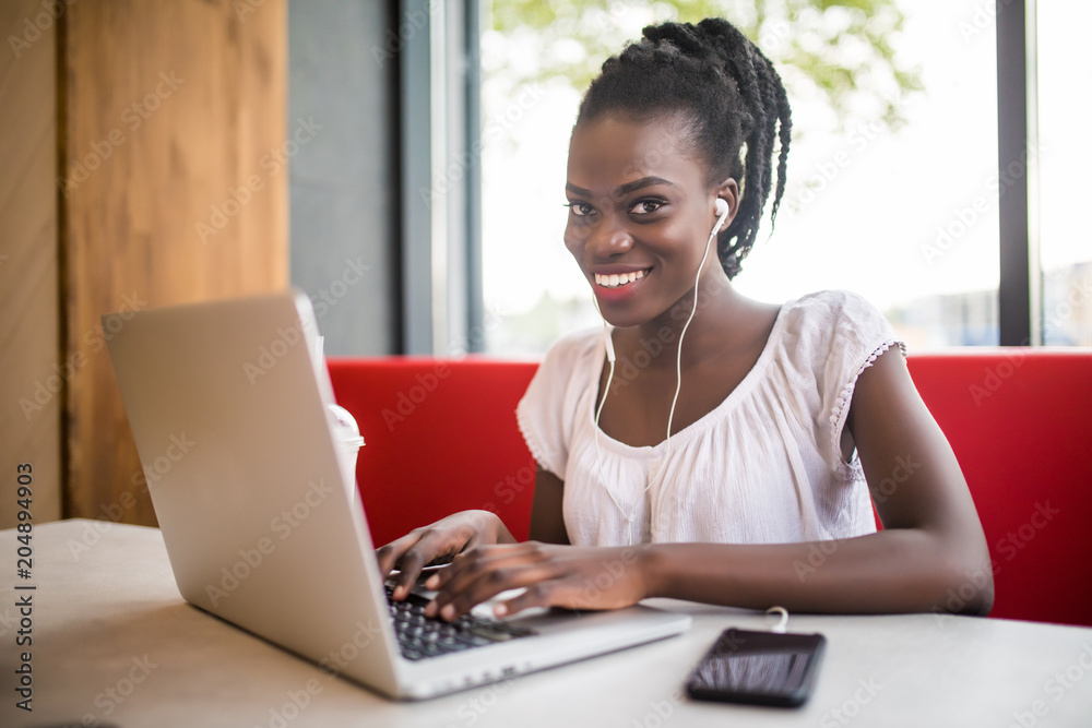 Cheerful female black model resting in coffee shop listening favorite music in headphones and chatting in networks. Beautiful dark skinned freelancer working on laptop Cheerful female black model resting in coffee shop listening favorite music in headphones and chatting in networks. Beautiful dark skinned freelancer working on laptop