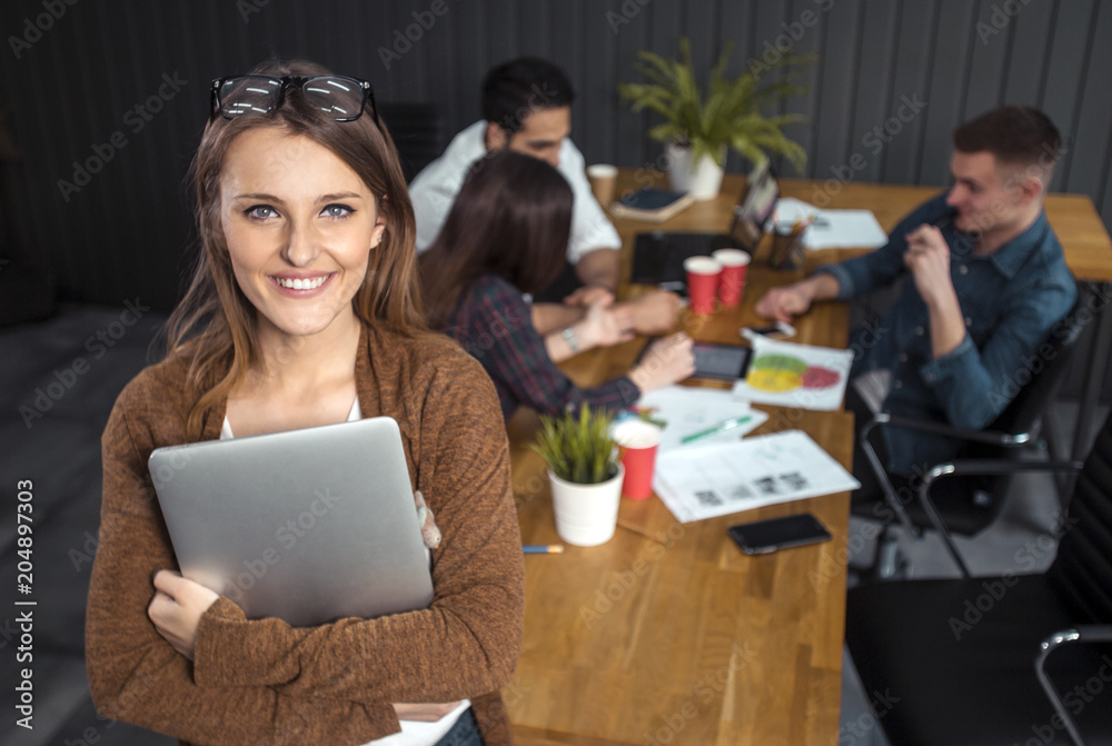 Pretty freelance woman standing with laptop in the office, three smart managers discussing possible solutions at the desk in the background, portrait shot Pretty freelance woman standing with laptop in the office, three smart managers discussing possible solutions at the desk in the background, portrait shot