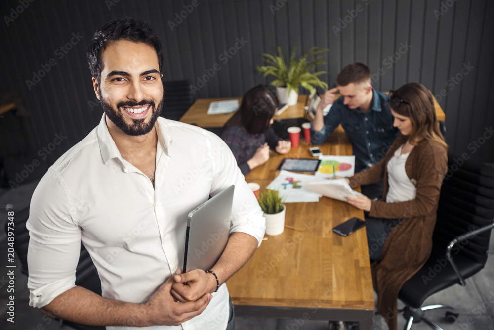 Fototapeta premium Pleased asian man with black beard holding silver laptop, standing before colleagues working on the task, preparing next stage of the project at office desk with graphs, coffee cups and gadgets