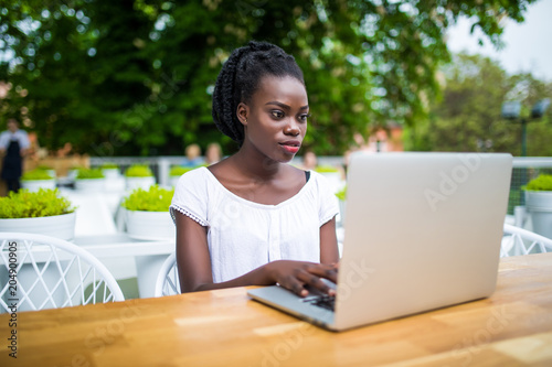 Beautiful young black female entrepreneur is sitting in street bar and working remotely on her project with laptop. Charming African American freelancer girl is sitting in cafe outdoor with netbook