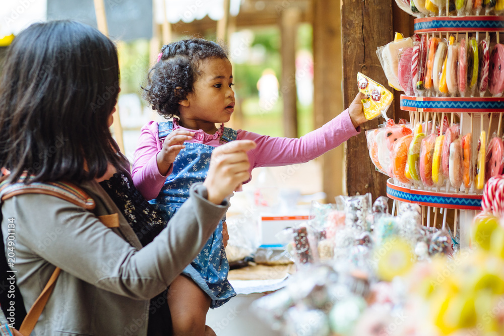 Mexican mother with dark skinned hispanic toddler girl choosing lollipop in candy showcase in open air market at summer time.