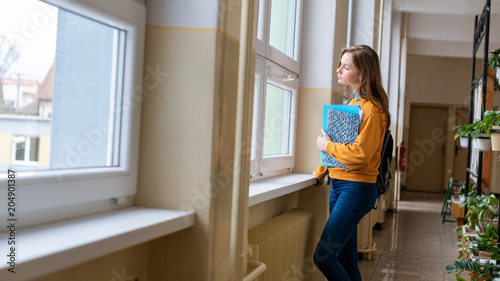 Young attractive female high school student standing by the window in the hallway at her school alone. Education concept. © andreaobzerova