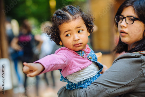 Children learning and family real life concept. Candid moment of latino american mother and dark skinned hispanic toddler daughter crying and pointing somewhere during a walk outdoors.