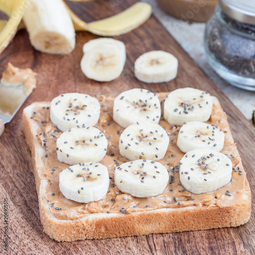 Healthy sandwich with crunchy peanut butter, banana and chia seeds, on wooden board, ingredients on background, horizontal, square format