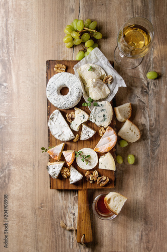 Cheese plate assortment of french cheese served with honey, walnuts, bread and grapes on rustic wooden serving board with glass of white wine over wood texture background. Top view, space.
