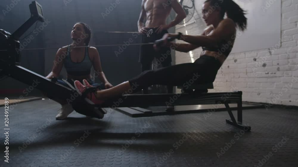 Fit woman working out on row machine in gym with trainers. Healthy young woman exercising cardio using rowing machine in fitness gym.