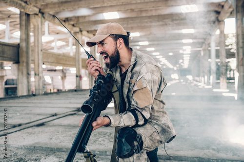 A picture of emotional and adult soldier screaming in portable radio. He is holding it in right hand. There is a big rifle in left one. Man wears military uniform. He is in hangar. photo