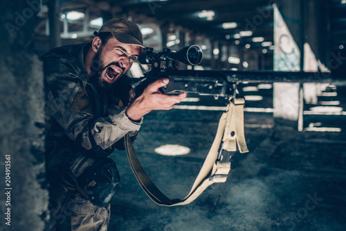 Horizontal picture of soldier is sitting on the ground on one knee near column and screaming. He is taking aim. Guy is using rifle for that. He is looking through rifle's lens. photo