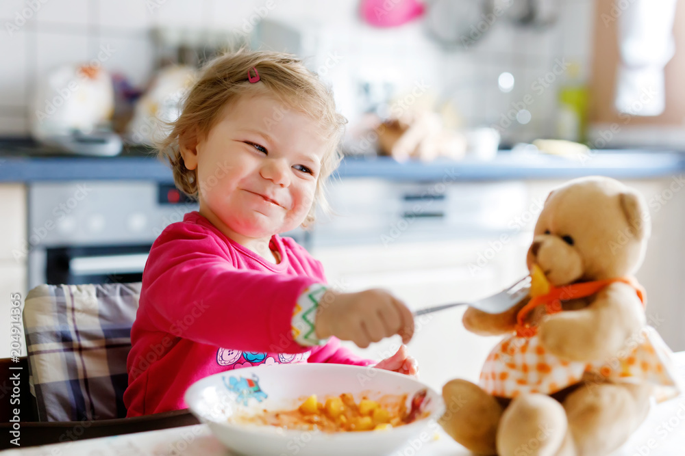 Adorable baby girl eating from fork vegetables and pasta. Little child feeding and playing with toy teddy bear. Adorable baby girl eating from fork vegetables and pasta. Little child feeding and playing with toy teddy bear.