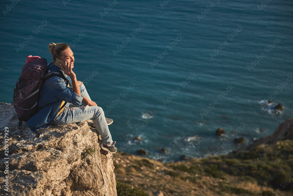 Glad female tourist communicating by cellphone while sitting on the top of mounting by the sea. Copy space in right side