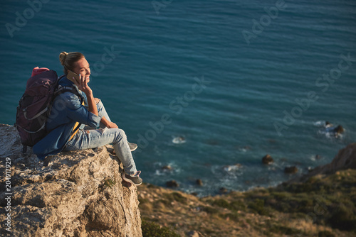 Glad female tourist communicating by cellphone while sitting on the top of mounting by the sea. Copy space in right side