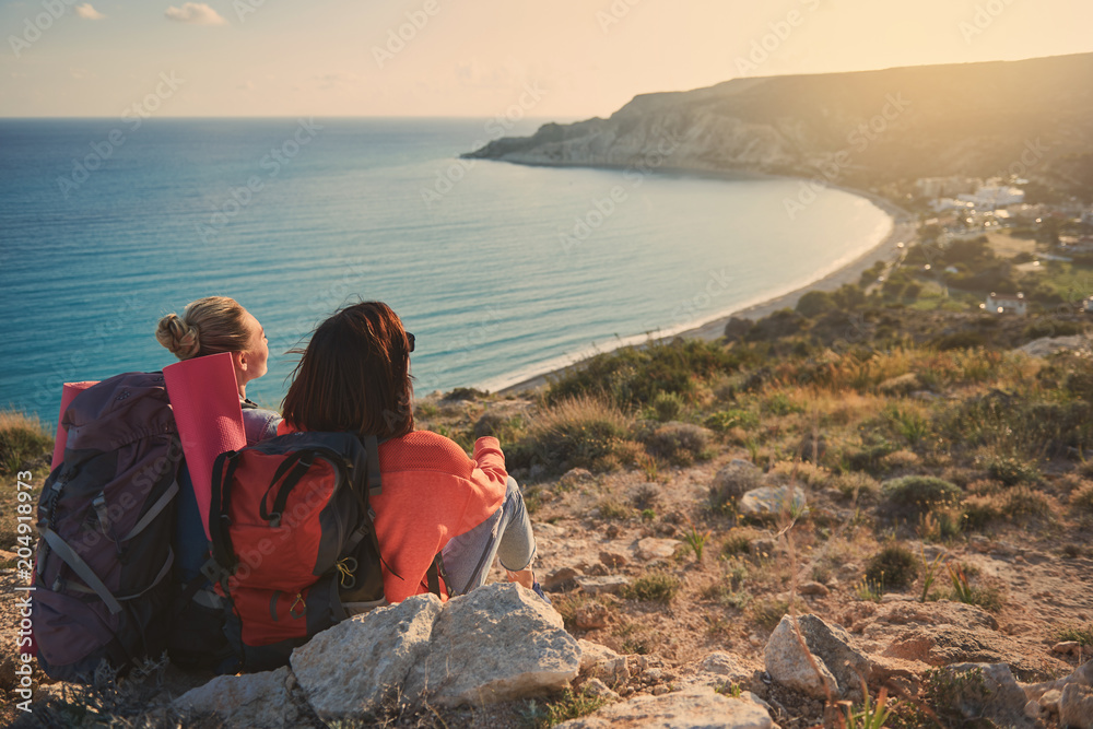 Two women with knapsacks sitting with their backs and looking at beautiful seaside. Copy space in right side Two women with knapsacks sitting with their backs and looking at beautiful seaside. Copy space in right side