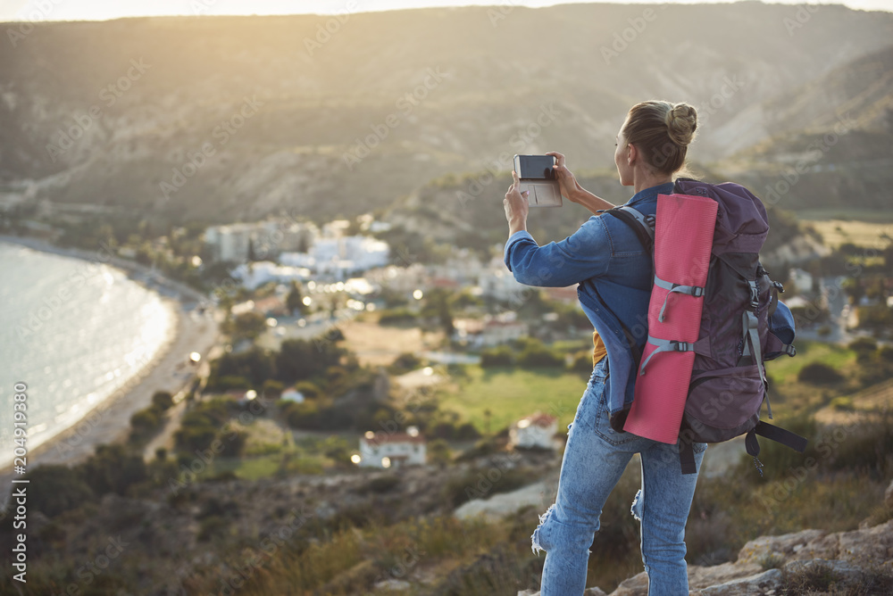 Focus on female person standing with her back and taking picture of coastline from high. She is carrying rucksack behind. Copy space in left side
