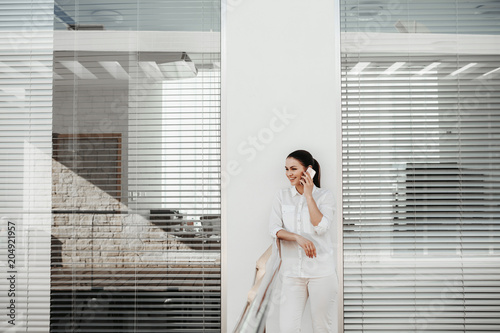 Portrait of laughing female person standing on balcony and using gadget for communication. Office blinds on background. Copy space in left side