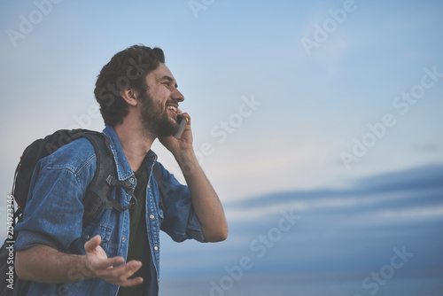 You will not believe where I am now. Happy young man is talking on mobile phone while hiking with backpack. He is looking at sea view with admiration and smiling