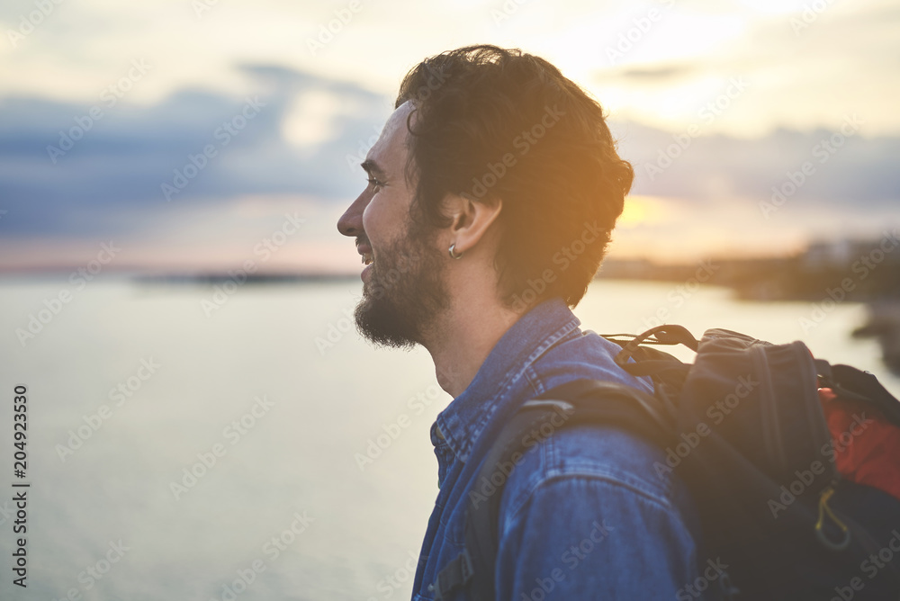 Profile of happy young man enjoying scenery at the seaside. He is standing with backpack and smiling Profile of happy young man enjoying scenery at the seaside. He is standing with backpack and smiling