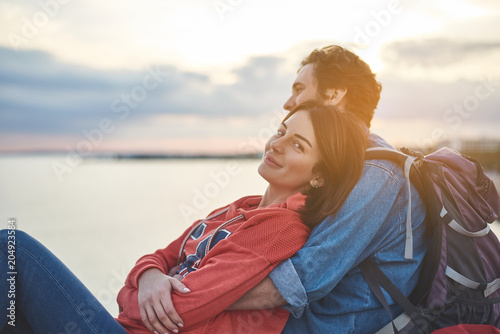 Portrait of satisfied girl enjoying nature together with her boyfriend. Couple is embracing while watching sunset at the seaside