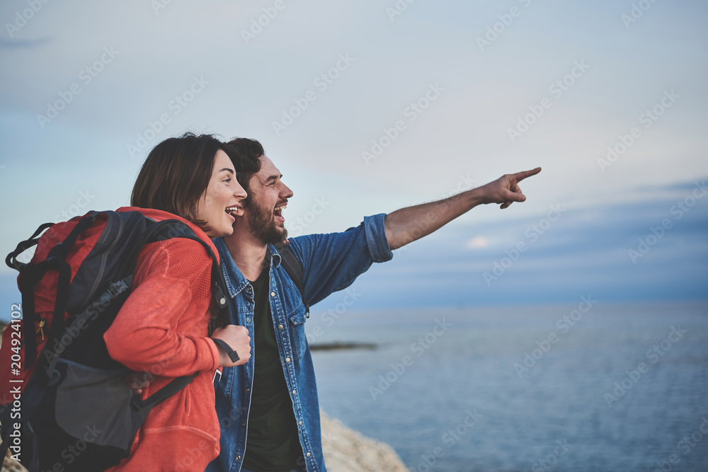 Look there. Side view of glad young man is pointing finger at horizon while embracing woman. Tourists are standing at the seaside Look there. Side view of glad young man is pointing finger at horizon while embracing woman. Tourists are standing at the seaside
