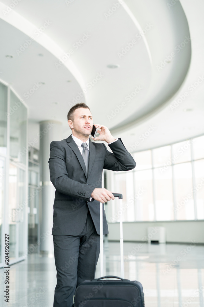Portrait of successful businessman standing in hall of modern airport with suitcase and speaking by phone Portrait of successful businessman standing in hall of modern airport with suitcase and speaking by phone