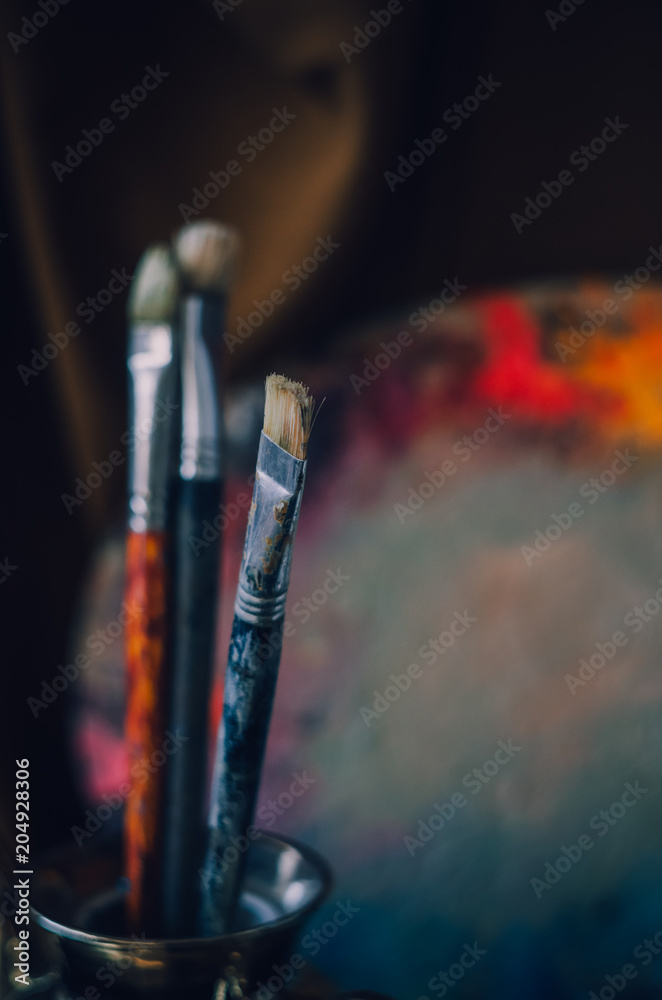 Naklejka premium Paintbrushes in the glass in a vertical position closeup. A colorful palette with oil paints on the background