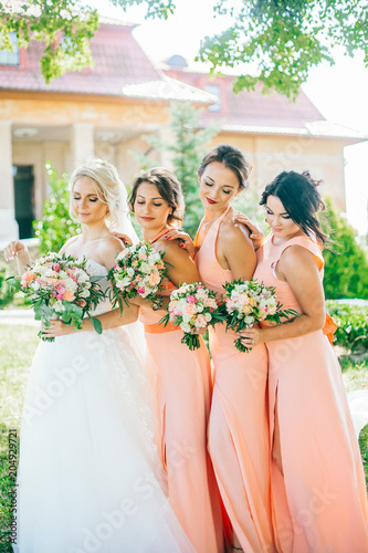 Stylish young smiling blonde bride with her bridesmaids in peach summer dress in the park on her happy wedding day. Women in the same color dress. All brunette one blonde. photo