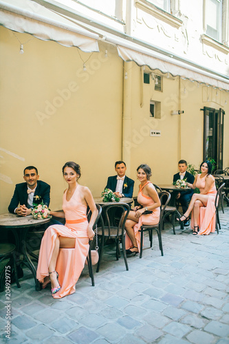 Happy fun newlyweds posing in street with bridesmaids & groomsmen. Bride with groom sitting at street cafe outdoor. 