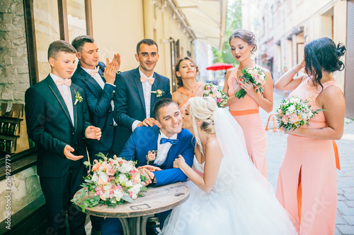 Happy fun newlyweds posing in street with bridesmaids & groomsmen. Bride with groom sitting at street cafe outdoor. 