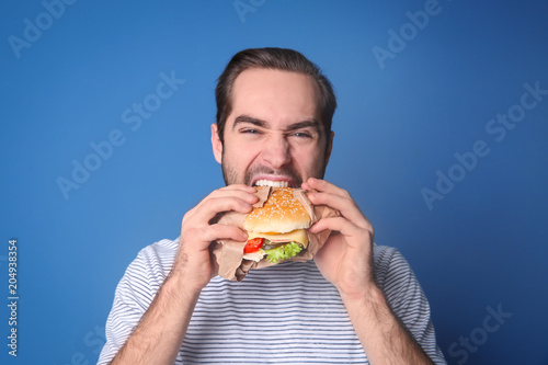 Handsome man eating yummy burger against color background