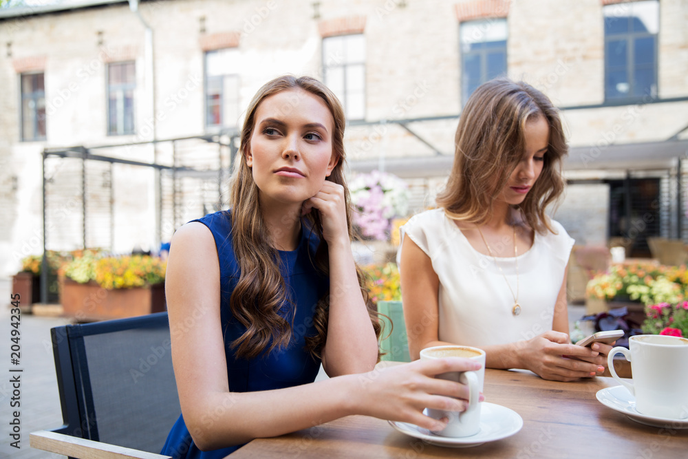 technology, internet addiction and people concept - young woman using smartphone and sad friend drinking coffee at cafe outdoors technology, internet addiction and people concept - young woman using smartphone and sad friend drinking coffee at cafe outdoors