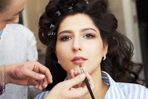 The make-up artist doing a makeup for bride. Makeup artist applies red lipstick . Beautiful woman face. Hand of make-up master, painting lips of young beauty model girl . Make up in process