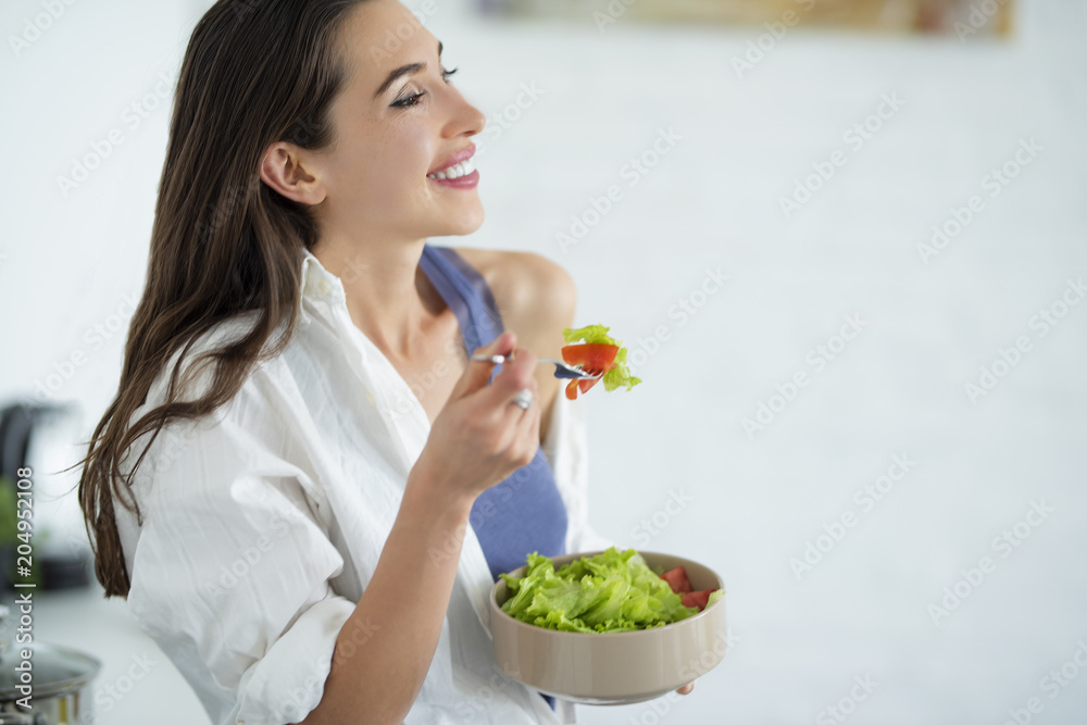 A beautiful young woman is cheerful while eating a salad, standing in her kitchen