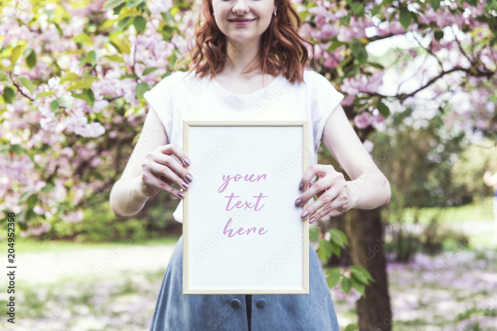 Young hipster redhead girl holding a photo frame in springtime blossom cherry trees garden. Creative mock up in the garden of cherry blossoms. Spring time. Young hipster redhead girl holding a photo frame in springtime blossom cherry trees garden. Creative mock up in the garden of cherry blossoms. Spring time.