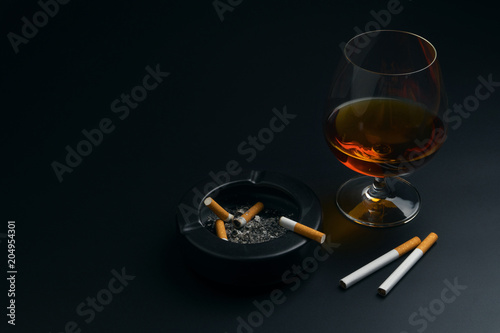 a glass of brandy, cigarettes and black ceramic ashtray full of ashes with the cigarette butt, on the black table