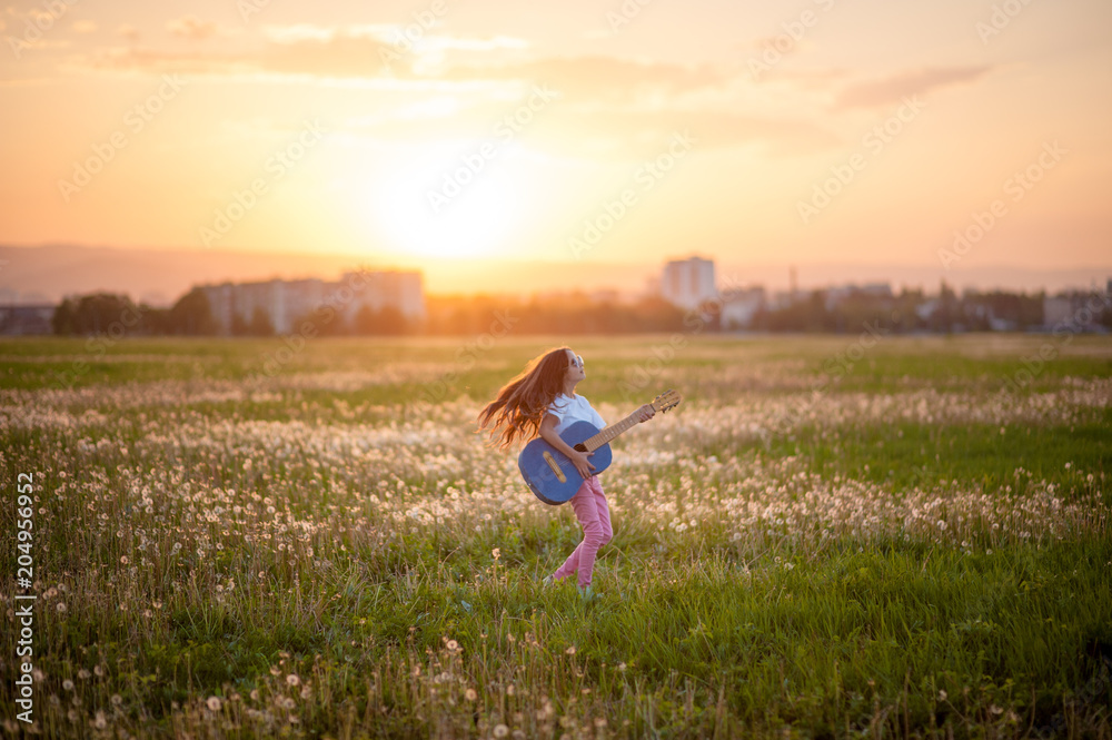 cute little girl with long hair and guitar in hands among blossom field in summer sunset having fun