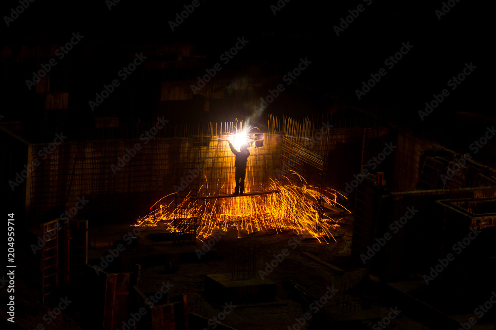 Obraz premium Building construction concept. Industry worker welding steel structure of building’s wall at night. Sparks flying from grinder.