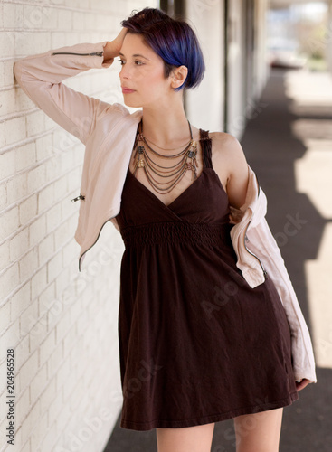 Woman in Tan Jacket, Brown Dress, and Necklace Leaning Against Wall Outside