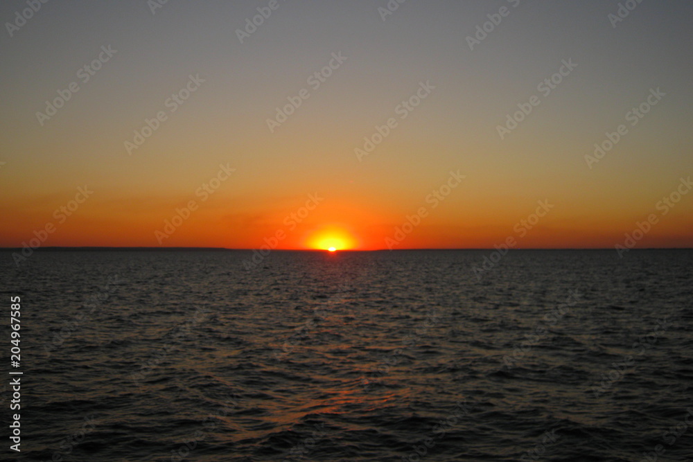 Beautiful orange sunset on the lake in summer against the setting sun on the horizon and the evening cloudless sky above the water Beautiful orange sunset on the lake in summer against the setting sun on the horizon and the evening cloudless sky above the water
