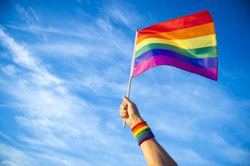 Colorful backlit rainbow gay pride flag being waved in the breeze against a sunset sky.