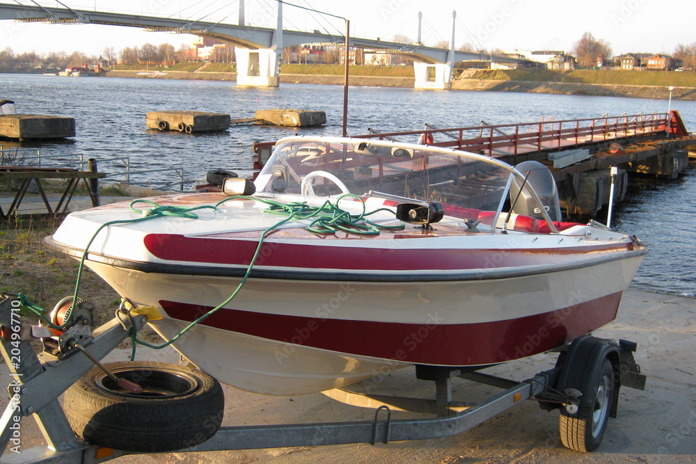 Descent boats on the water – red and white boat with an outboard motor on the trailer near the water fall at the background of the river, Wharf and bridge