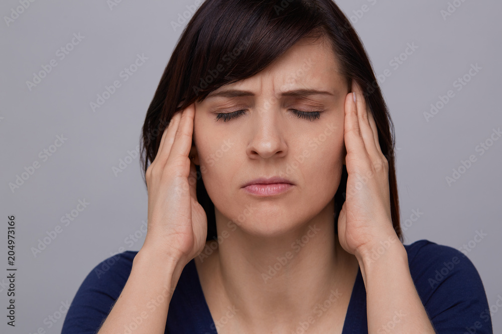 Closeup portrait of young woman on grey background suffering from strong headache, holding fingers to temples and closing eyes from pain.Severe migraine. Closeup portrait of young woman on grey background suffering from strong headache, holding fingers to temples and closing eyes from pain.Severe migraine.