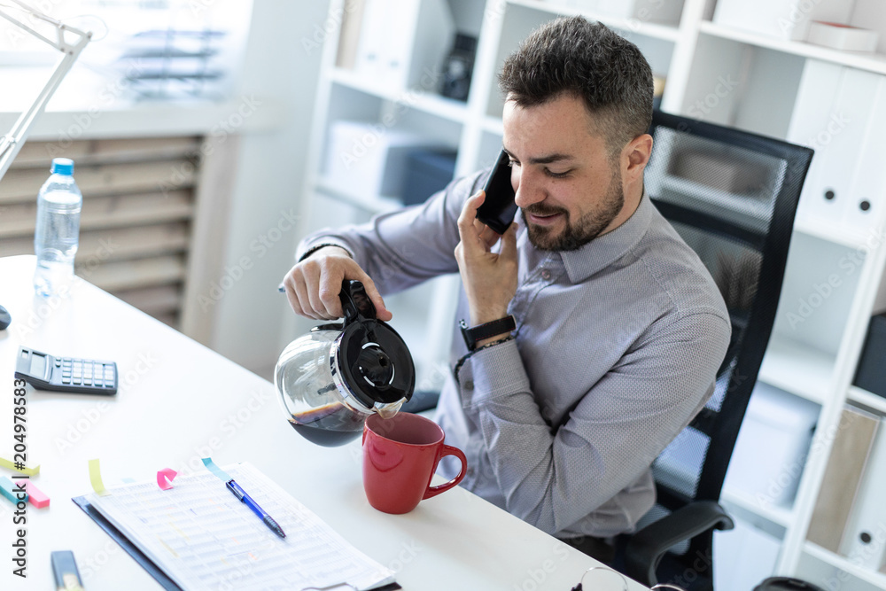 A young man in the office sits at a table, talking on the phone and pouring coffee into a cup. A young man in the office sits at a table, talking on the phone and pouring coffee into a cup.